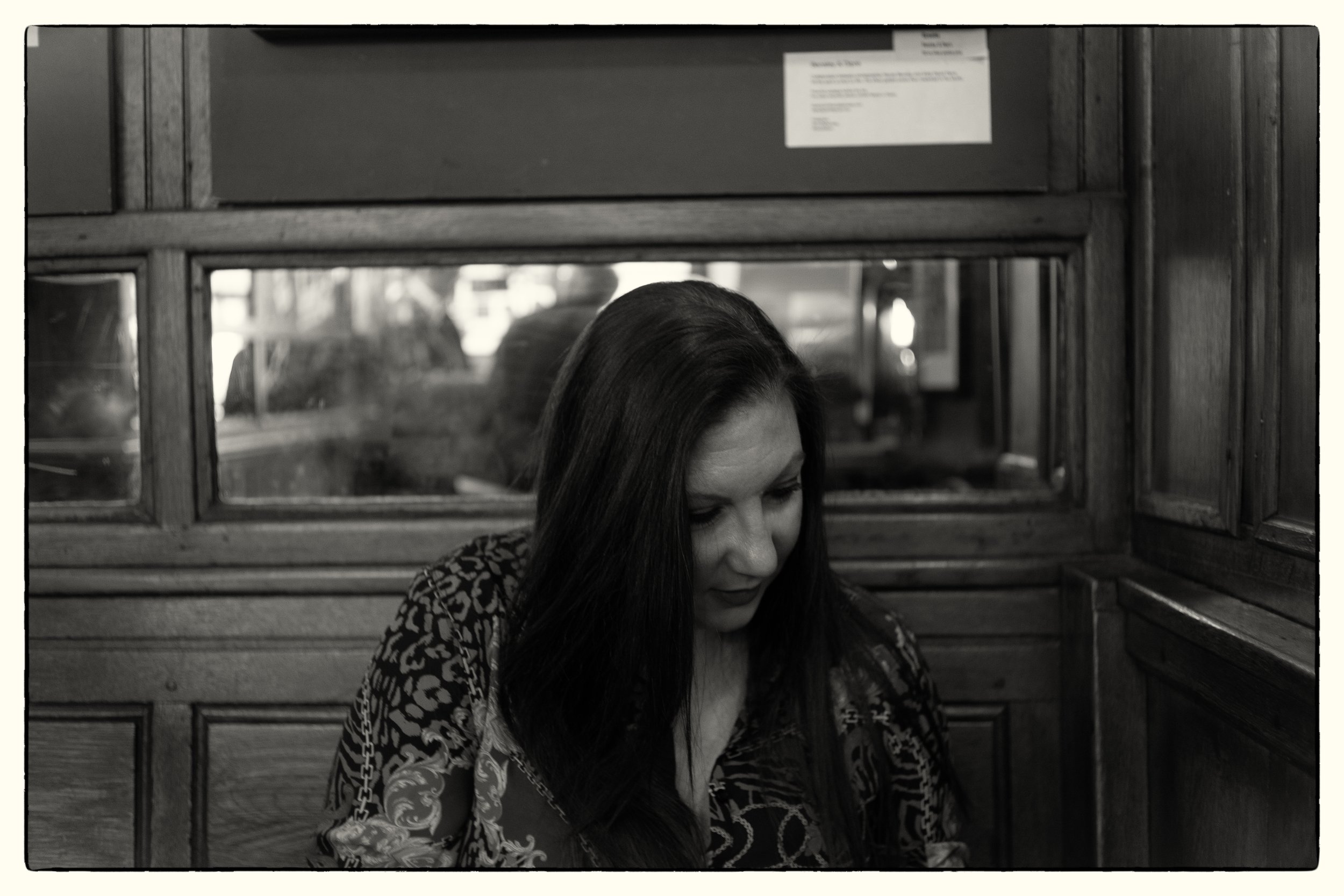 A woman with long dark hair wearing a patterned top, sitting in front of wooden paneling and a window, with a reflection in the glass behind her.