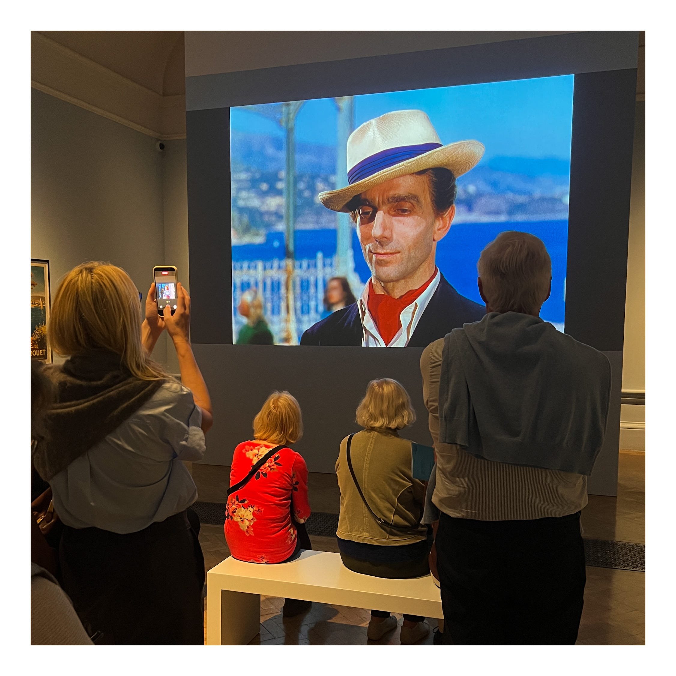 People gathered in a room watching a large screen displaying a historically iconic image of Fred Rogers, the beloved television personality, with a background of blue water and a fence, wearing a hat and suit.