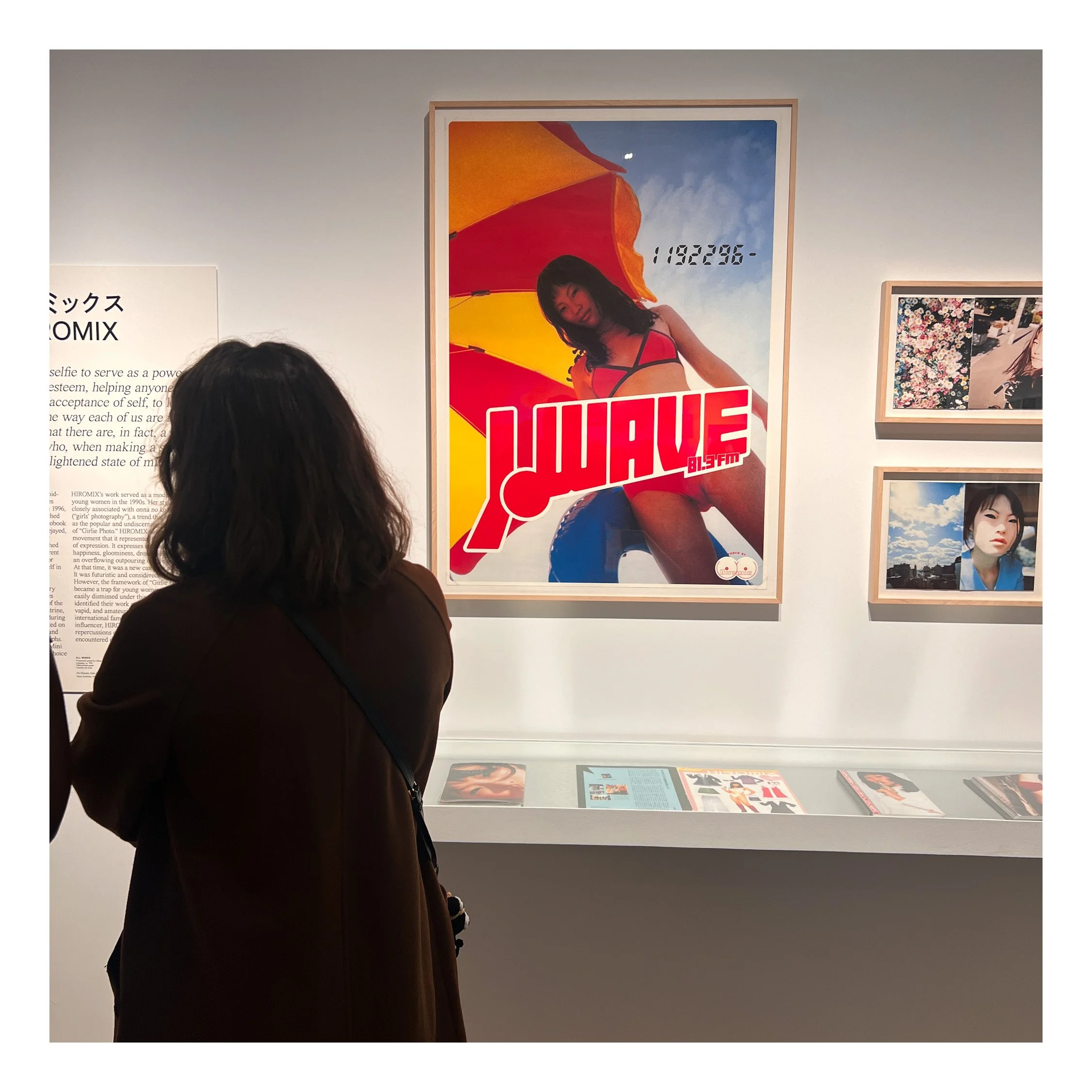 A person with dark hair and a brown coat observing artwork in a gallery, with framed pictures hanging on the white wall behind.