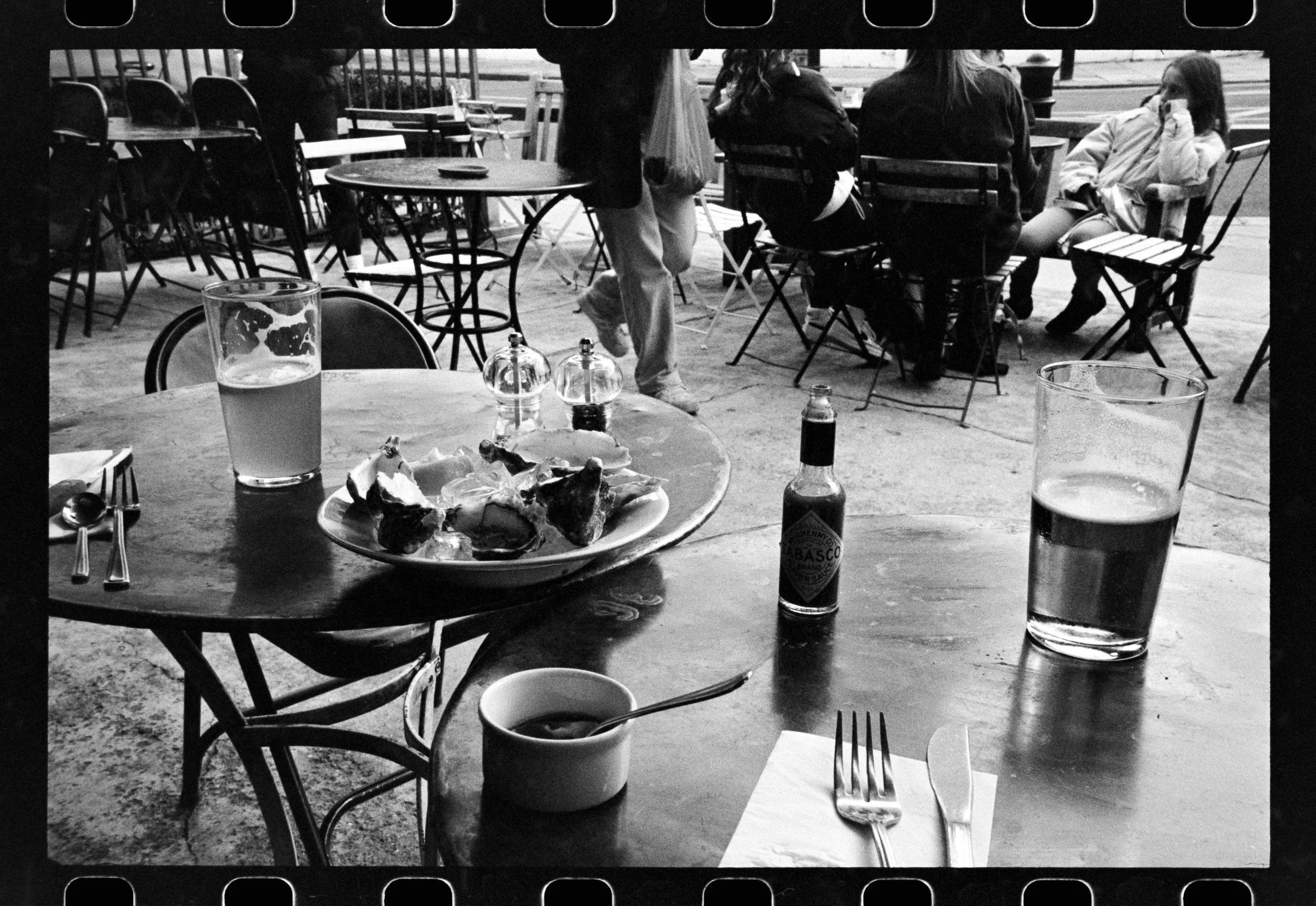 Black and white photo of an outdoor cafe or restaurant with empty tables and chairs. In the foreground, there is a table with a pizza plate, two glasses of beer, and condiment bottles. In the background, people are seated and walking, with some weari