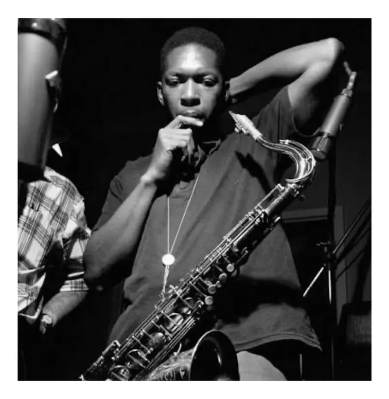 Black and white photo of a young man holding a saxophone, pondering with his hand near his face, in a music studio setting.