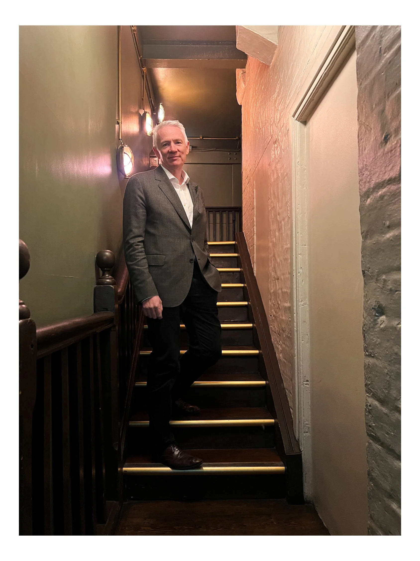 A man wearing a gray blazer and white shirt standing on a staircase indoors, with a dark colored wall with lighting on the left side and a textured gray wall on the right side.