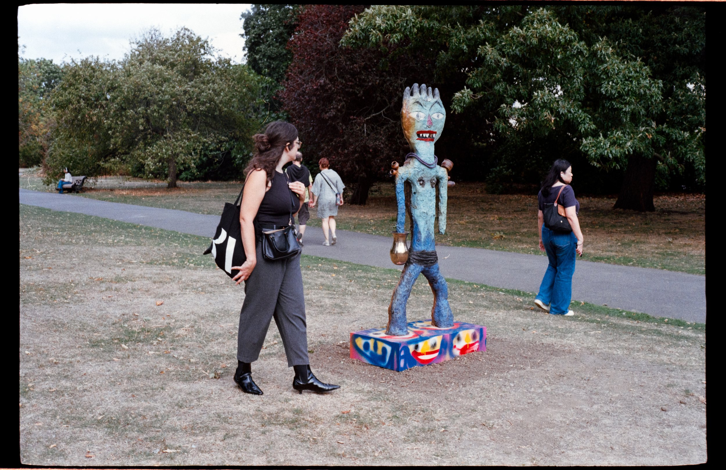 A woman in a black sleeveless top and gray pants stands on a park pathway, looking at a colorful, abstract sculpture of a creature with a large head, sharp teeth, and elongated limbs. The sculpture has a vibrant, graffiti-style base. Other park visit