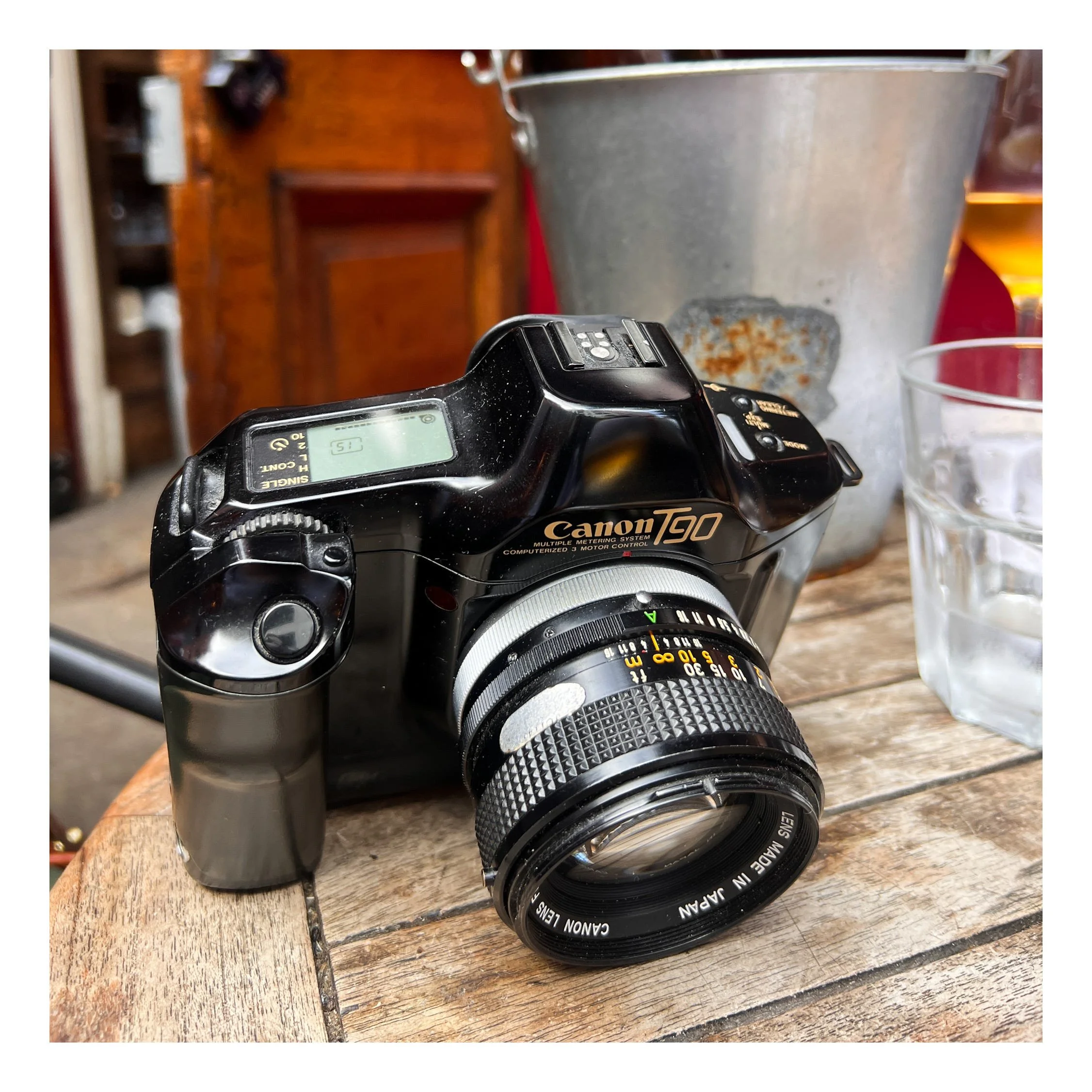 A Canon T90 camera with a zoom lens, a metal bucket, and a glass of water on a wooden surface.