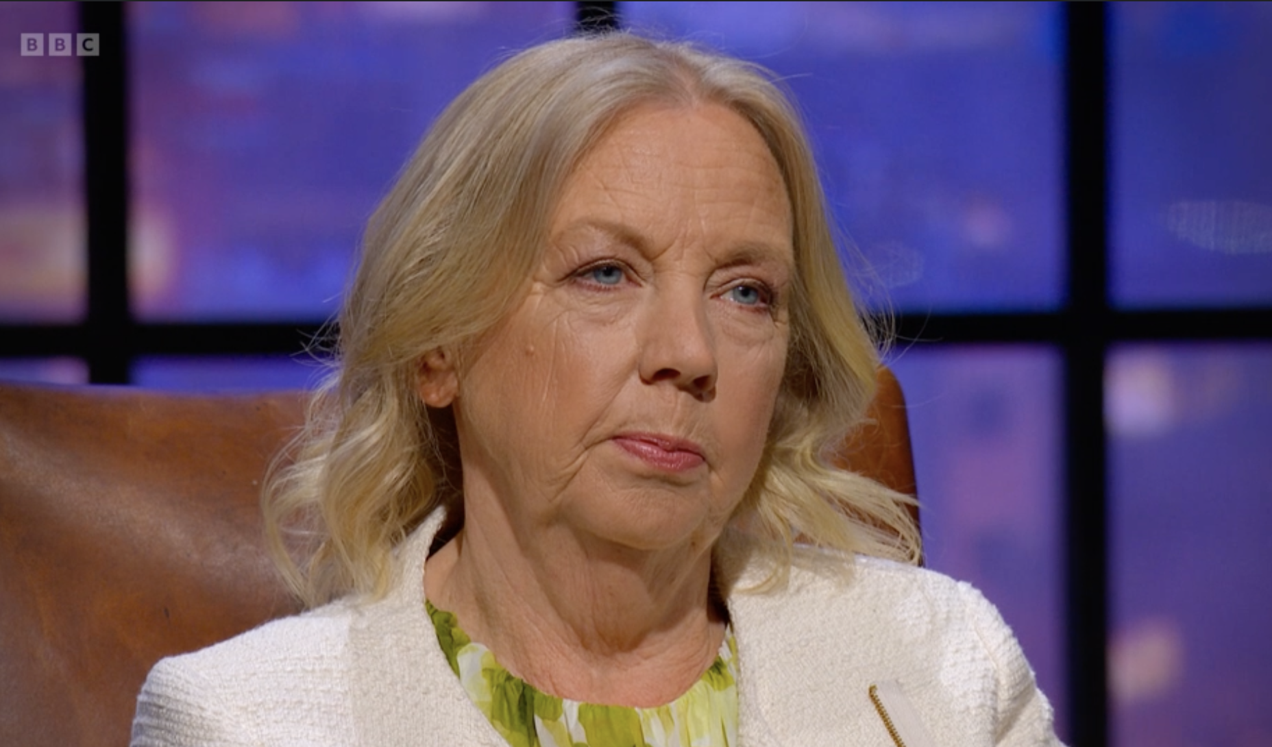 Close-up of a serious older woman with blonde hair, wearing a white jacket and green patterned blouse, sitting in a brown leather chair on a television interview set with purple and black background.