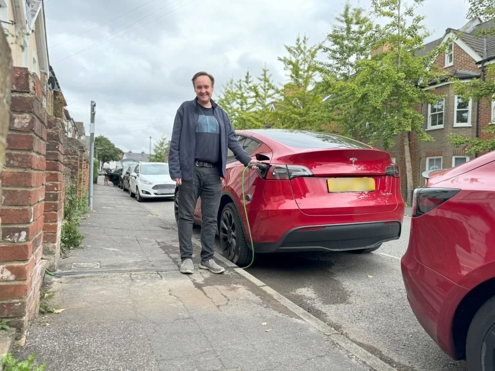 Man in black jeans and blue jacket stood on paveent charging a red car parked on the street