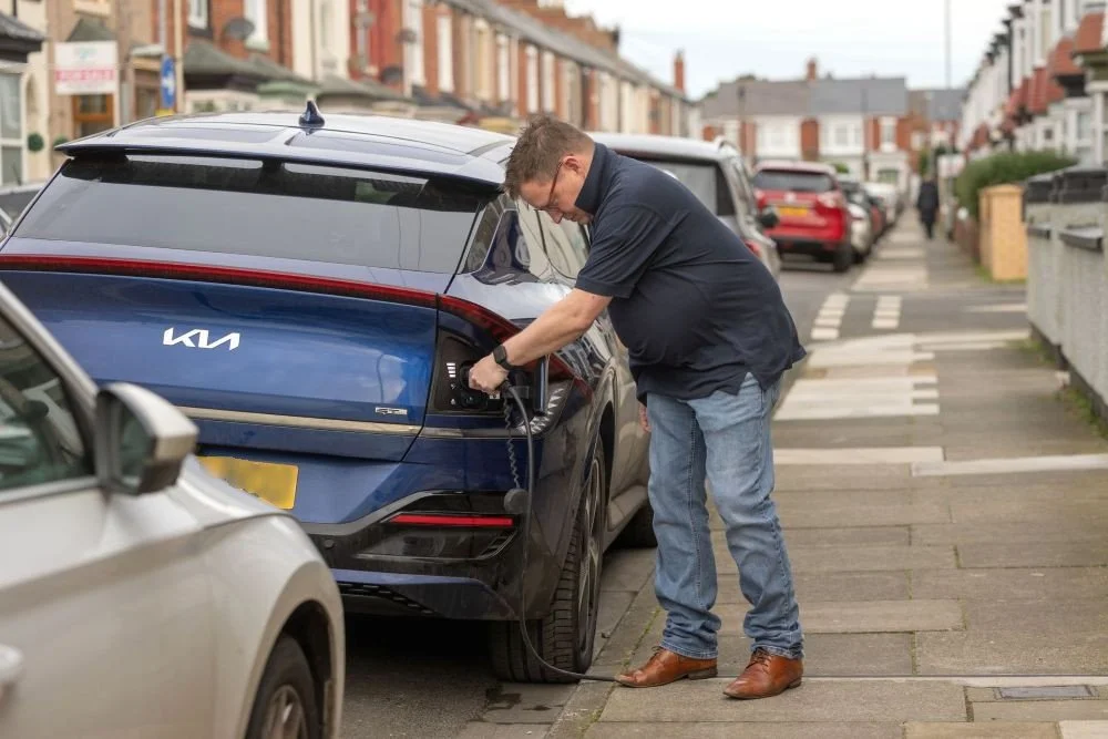 Man in blue jeans, brown shoes and, navy tshirt charging car parked on terraced street