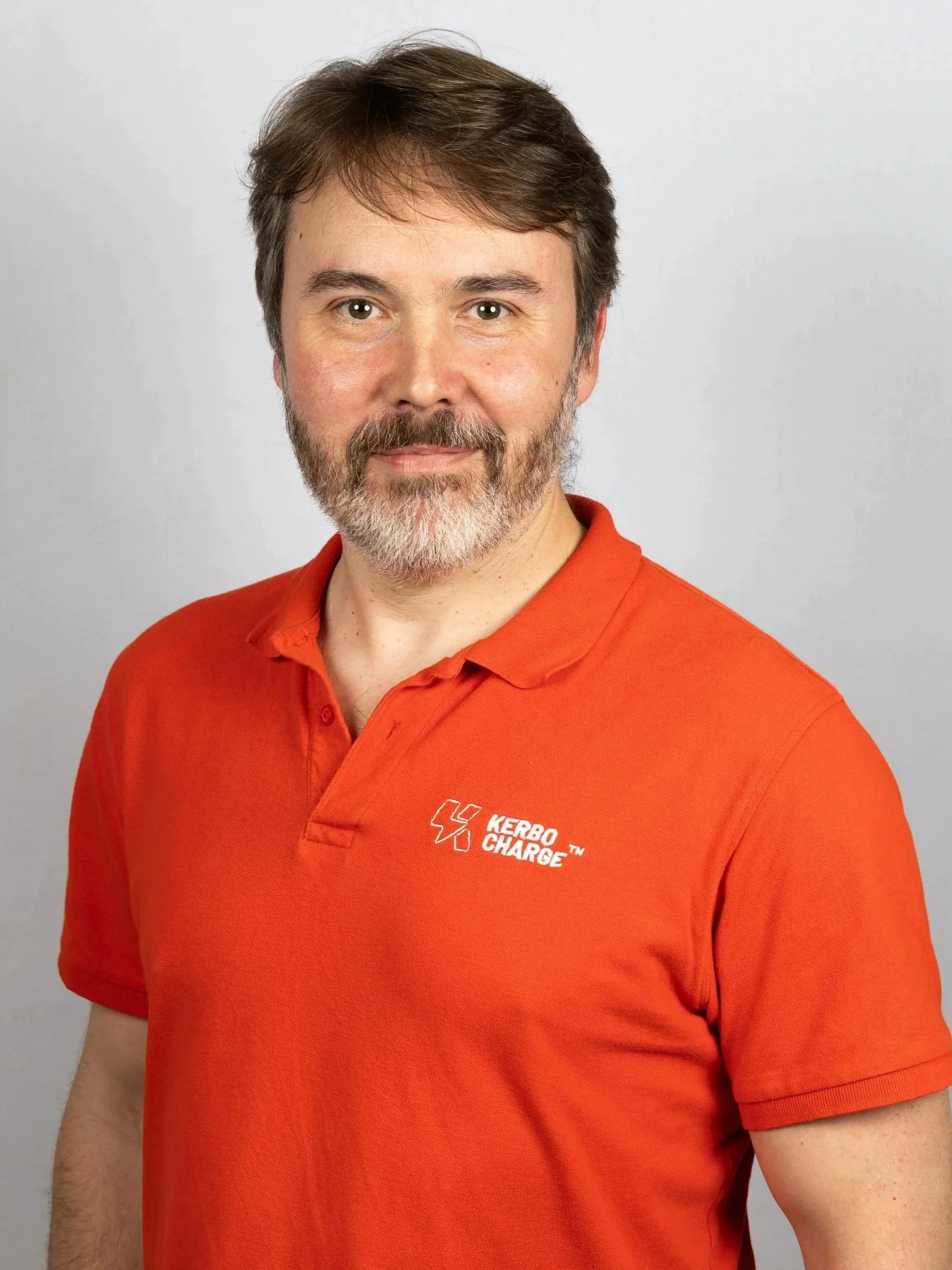 A man with brown hair, a beard, and a mustache, wearing an orange polo shirt with a logo that says 'KERBO CHARGE'. He is standing against a plain, light-colored background, looking directly at the camera with a slight smile.