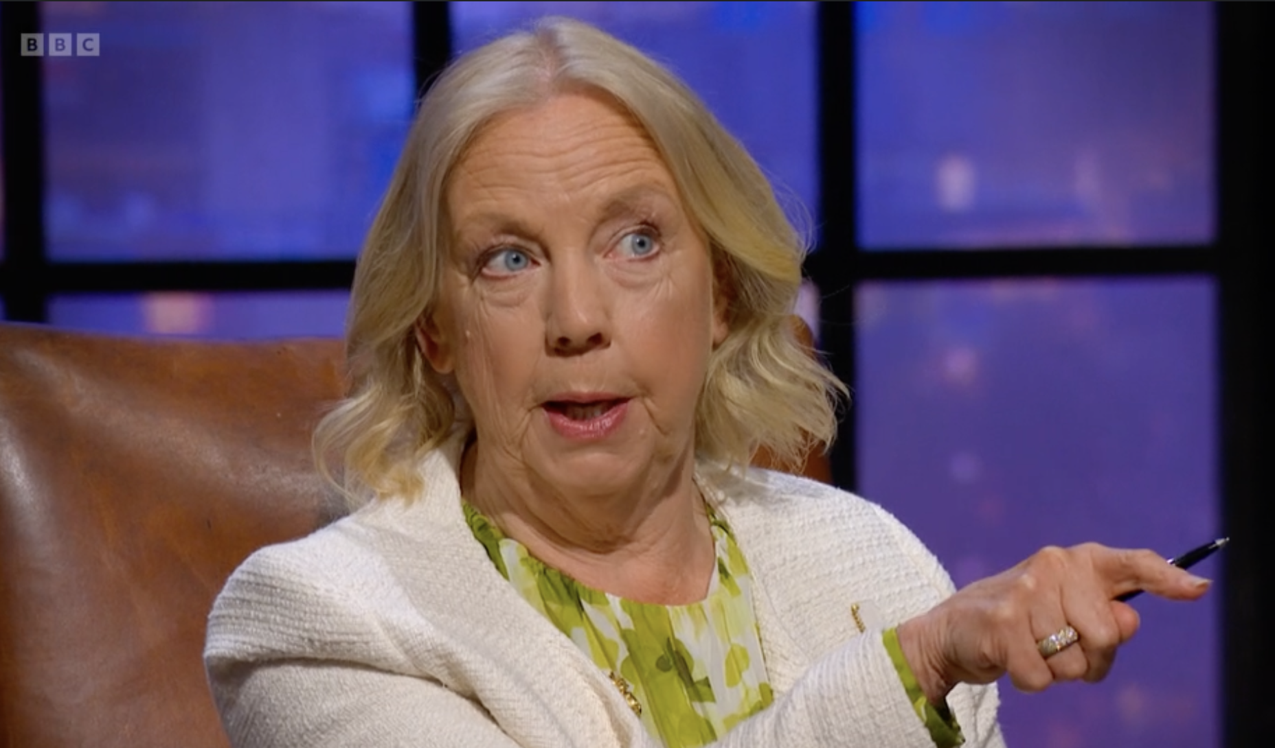 Older woman with blonde hair and blue eyes sitting in a room with purple screening in the background, wearing a white sweater over a green and yellow patterned top, holding a pen and gesturing with her hand.