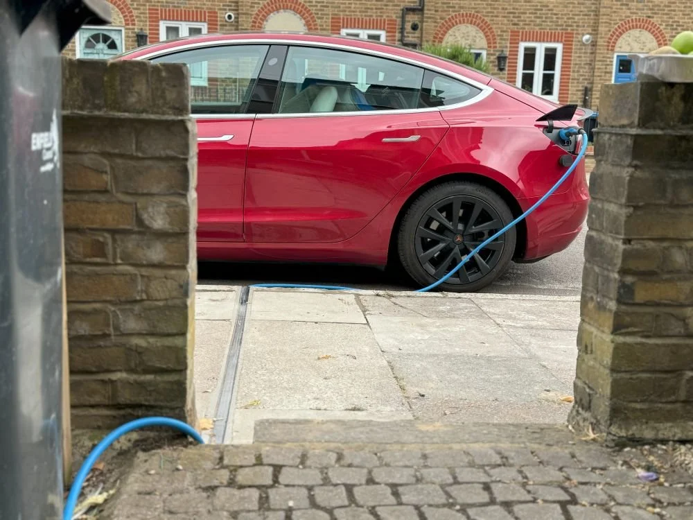 Red Tesla electric car charging at a sidewalk charging station, viewed through a brick gate.