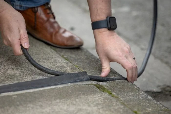 A person is placing EV cable charger into cross pavement cable gully inpavement, wearing brown leather shoes, a black smartwatch, and a silver ring.