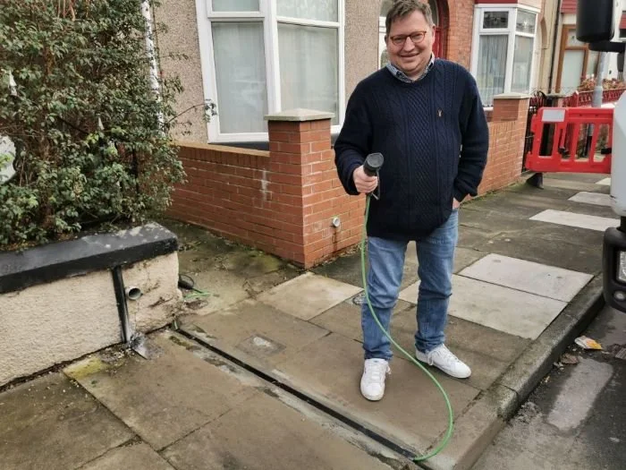 A man standing on a pavement an EV charging cable, wearing glasses, a navy sweater, jeans, and white sneakers, with a smile on his face. He is in front of a residential building with a brick wall and garden bush.