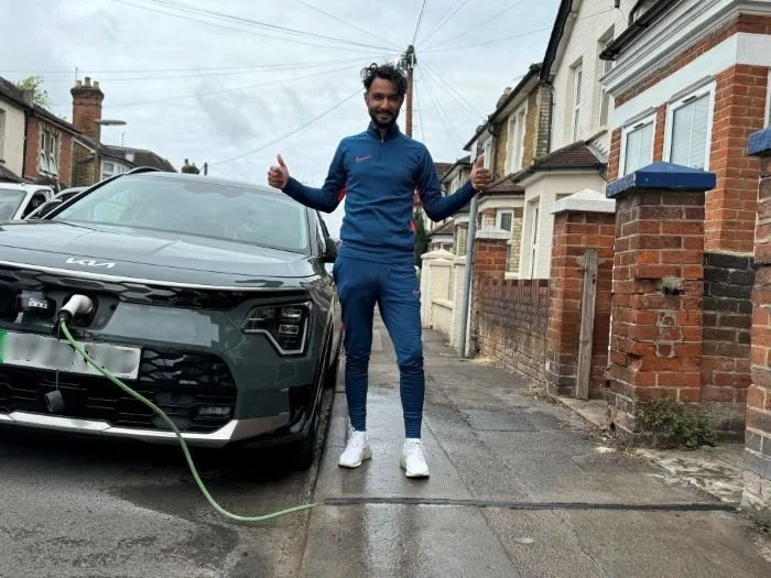 Man in blue tracksuit with two thumbs up stood next to EV being charged on the street