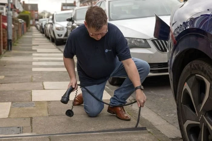 A man kneeling on a sidewalk, plugging in an electric vehicle charger into a car with a row of parked cars behind him.