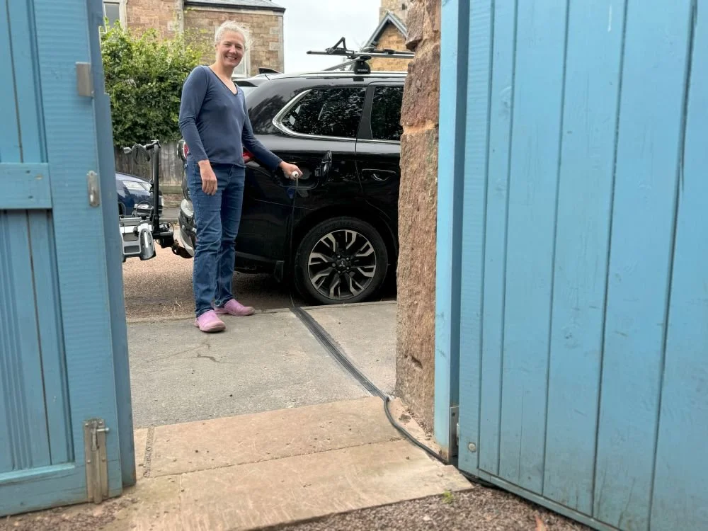 lady in jeans and blue jumper charging car on the street beyond the property gate
