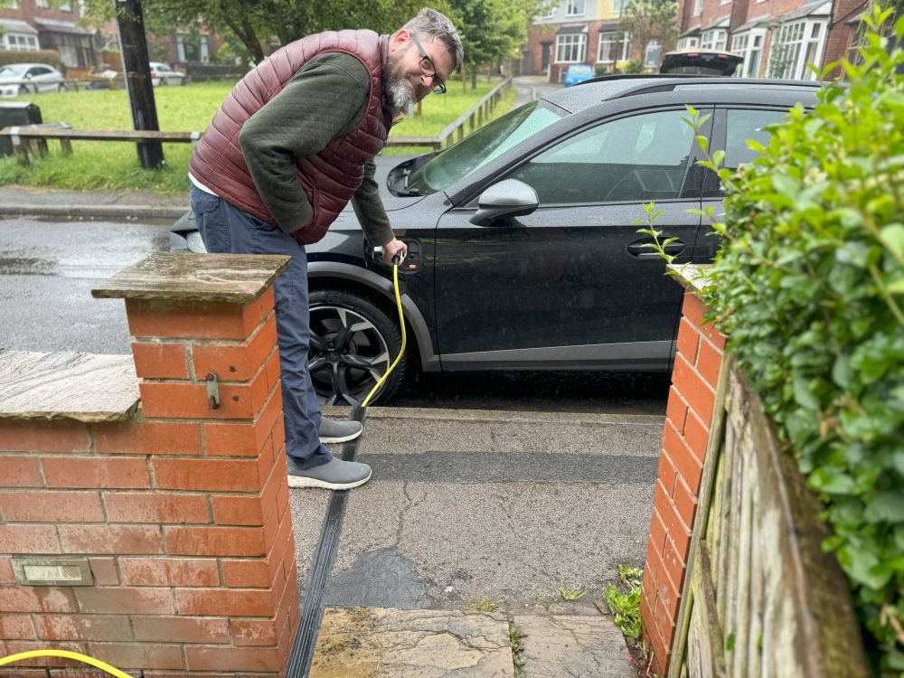Man in blue trousers, jumper and gilet bend down charging EV