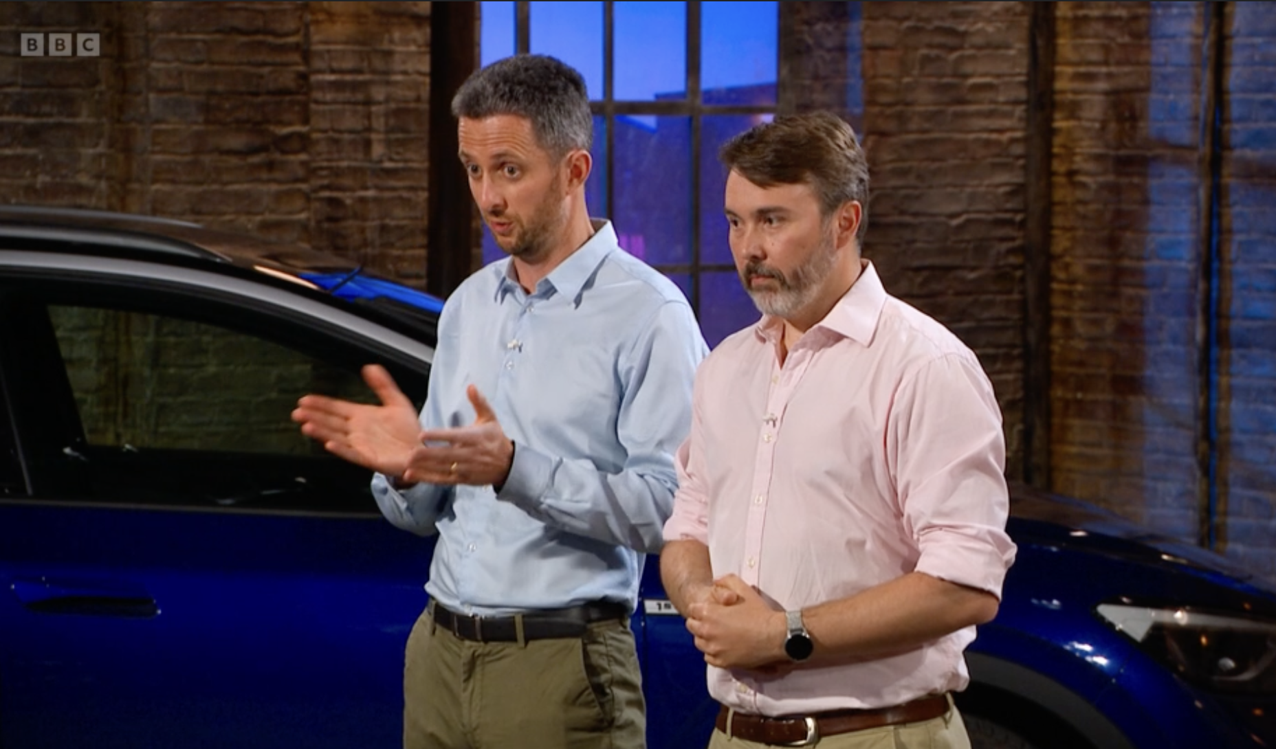 Two men standing in front of a blue car inside a brick-walled industrial-style studio.