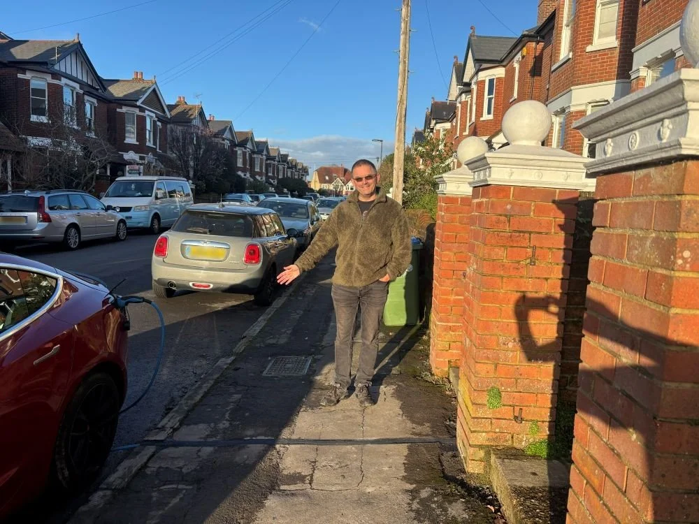 man in brown trousers and coat stood on pavement pointing to car being charged on the street
