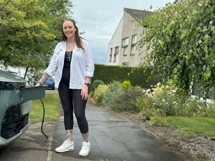 A woman with tattoos on her legs and arm charging an electric car in a residential neighborhood on a cloudy day.