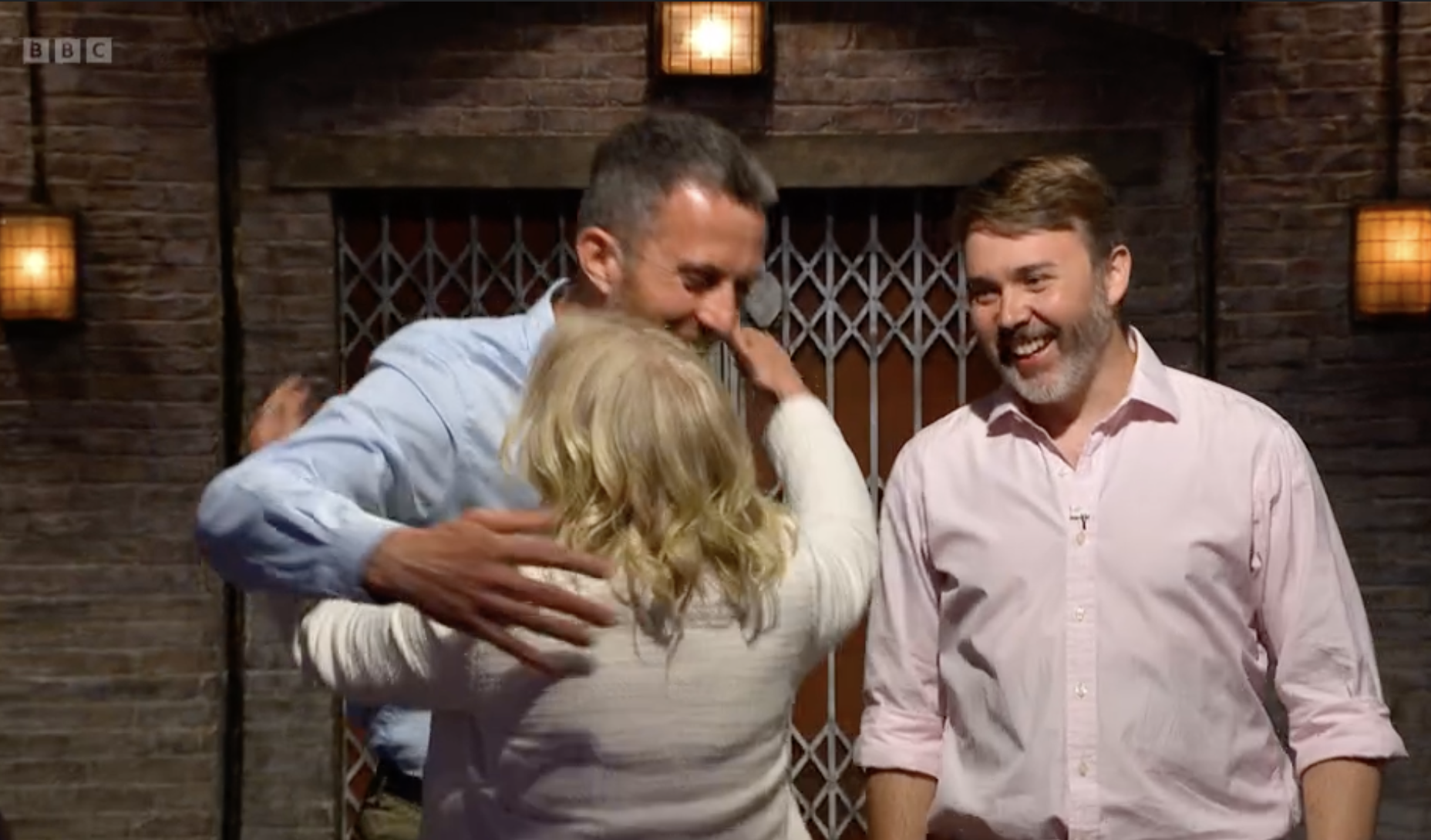 Three people sharing a group hug, smiling and embracing each other in a warmly lit indoor setting with brick walls and wall-mounted lights.