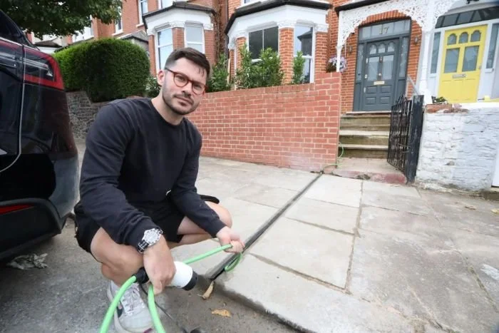Man squatting on sidewalk holding a garden hose, with brick houses and stairs in the background.