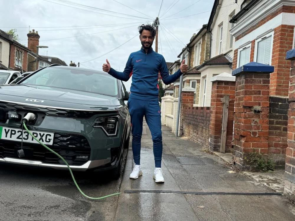 Man in blue tracksuit with two thumbs up stood next to EV being charged on the street