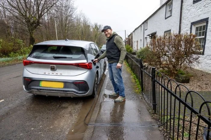 Man in jeans, hoodie and baseball cap charging a silver car parked on the side of the road