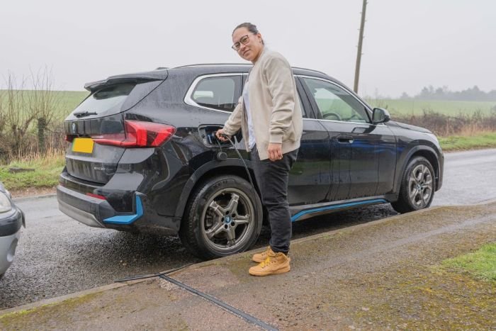 A person charging an electric car parked on the side of a wet, rural road on a cloudy day.