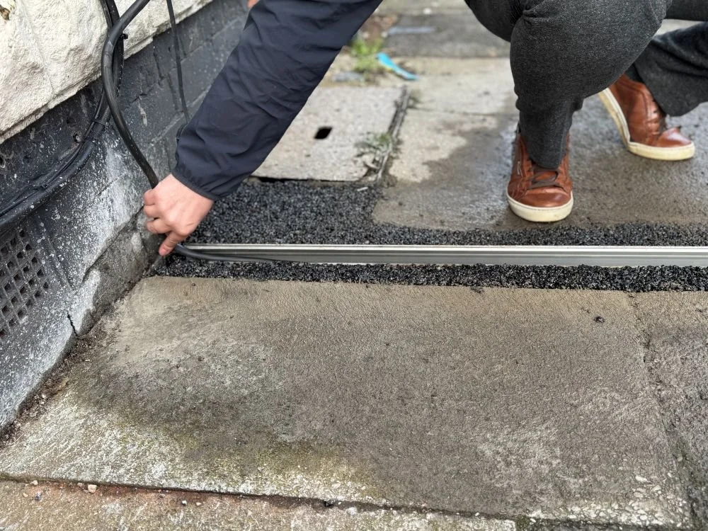 Person in brown trainers an dblue coat bending down to place EV charger cable into pavement gully