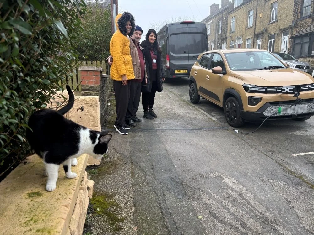 Cat stood on wall and 3 people stood on pavement next to charging EV parked on residential street