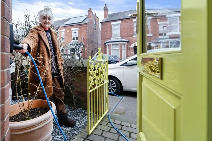 A woman with grey hair, wearing a brown coat, waters a plant outside her house, which has a yellow door and a yellow gate. A white car is parked in the street behind her, with brick houses in the background.
