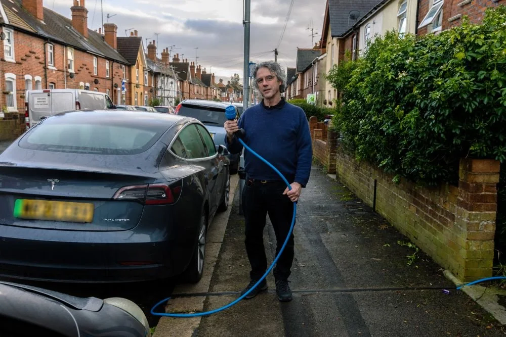 Man in black jeans and blue jumper stoof on pavement next to parked car holding an EV charging cable