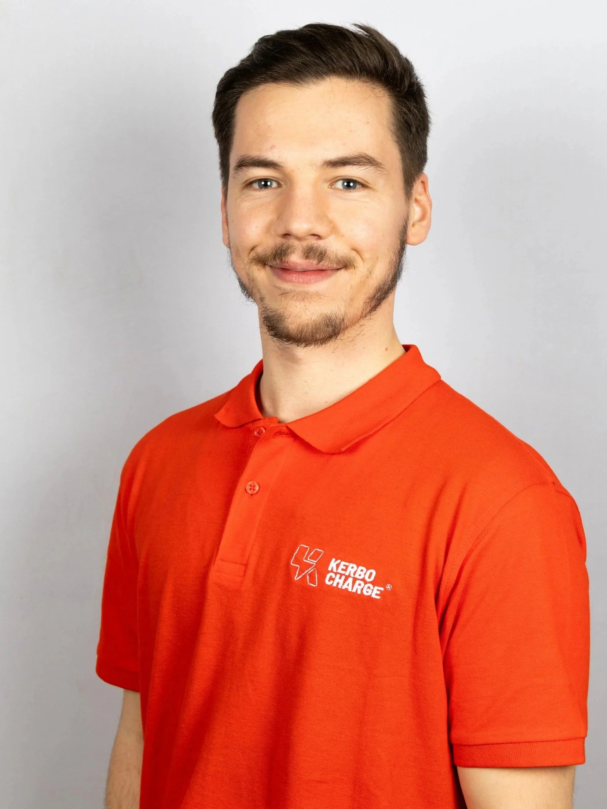 A young man with light skin, brown hair, and a short beard, smiling, wearing a red polo shirt with the 'Kerbo Charge' logo, standing against a plain light gray background.