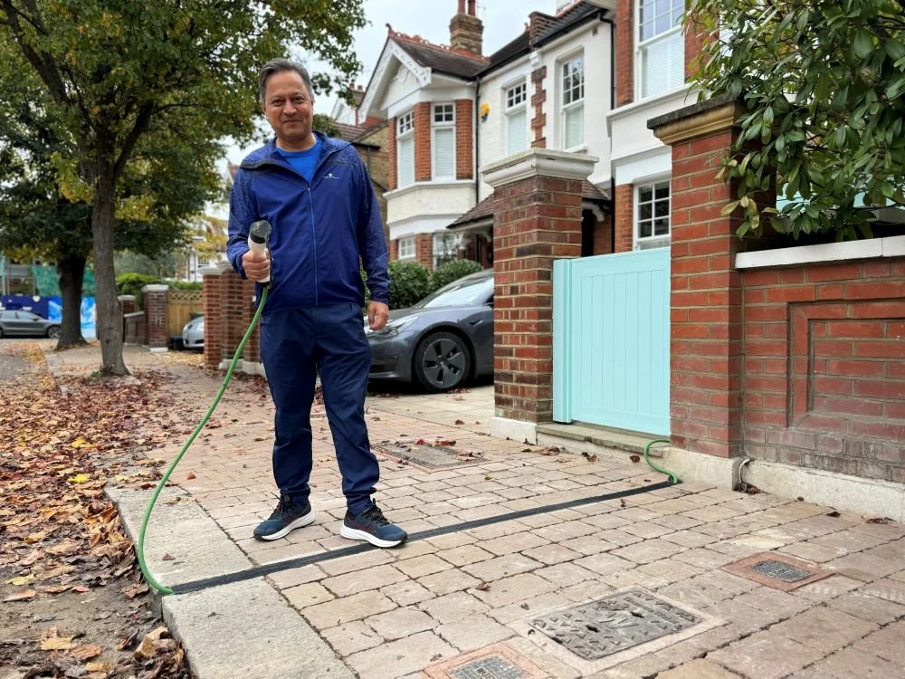 Man in blue tracksuit and trainers stood on pavement holding EV cable charger which is running through a pavement gully
