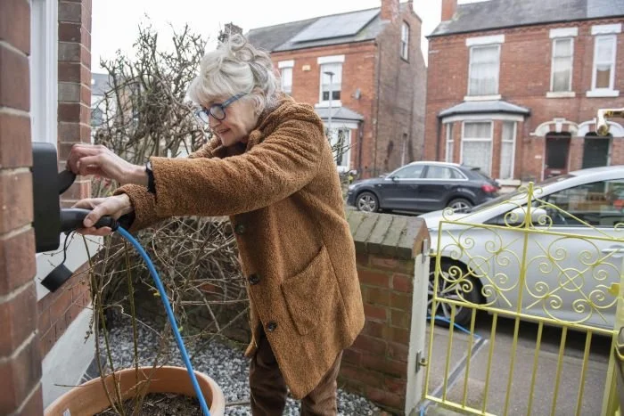 An elderly woman with gray hair, glasses, and a brown coat, is watering a plant outside her house with a blue hose.