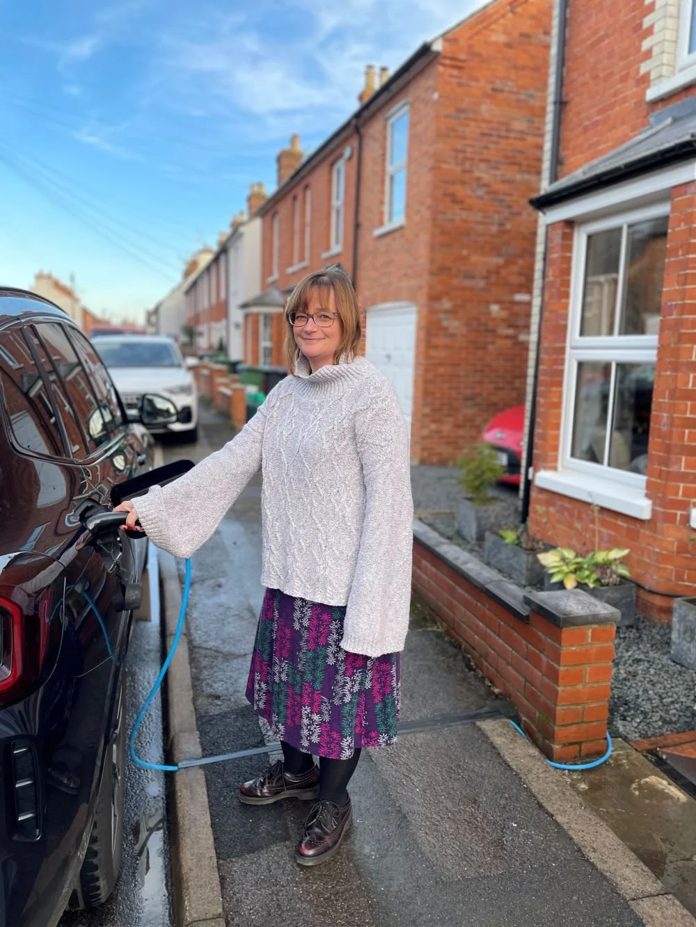 lady in skirt and jumper charging car on street outside terraced house
