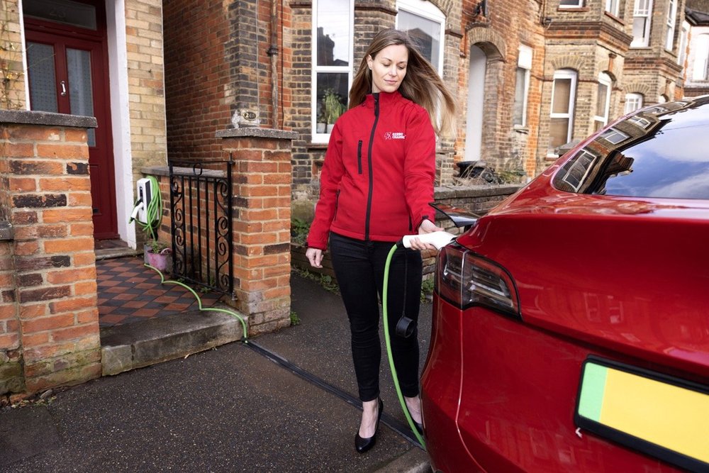 Woman in red jacket charging a red electric car using a charging station outside her home.