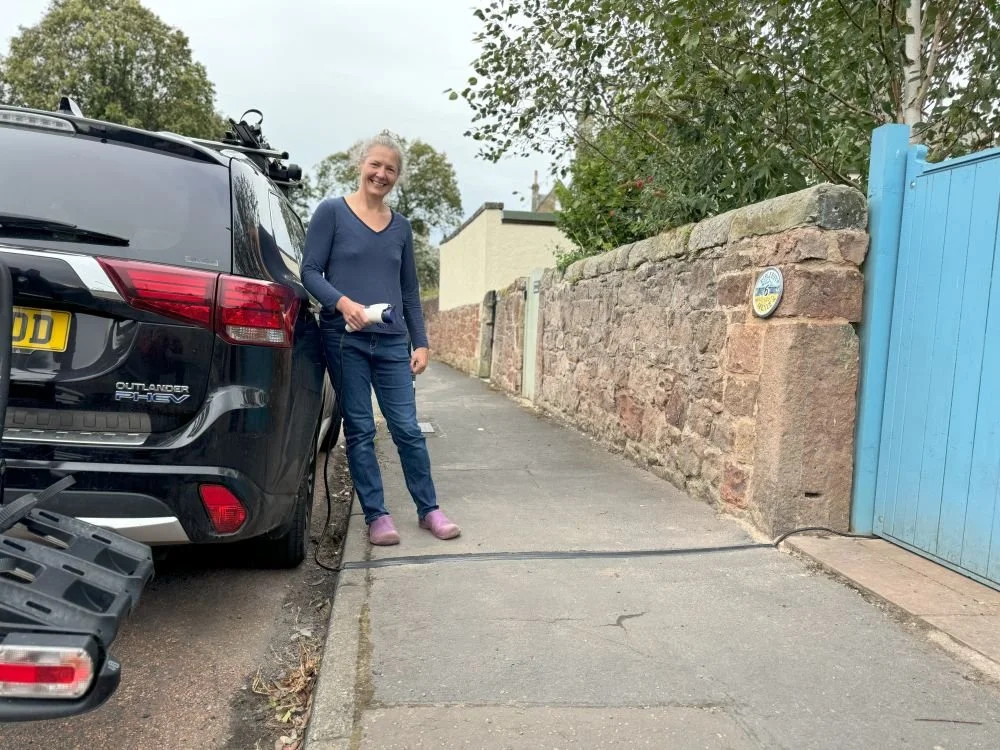 lady in purple crocs, blue jeans and jumper holding EV charger cable while leaning against a car parked on the side of the road