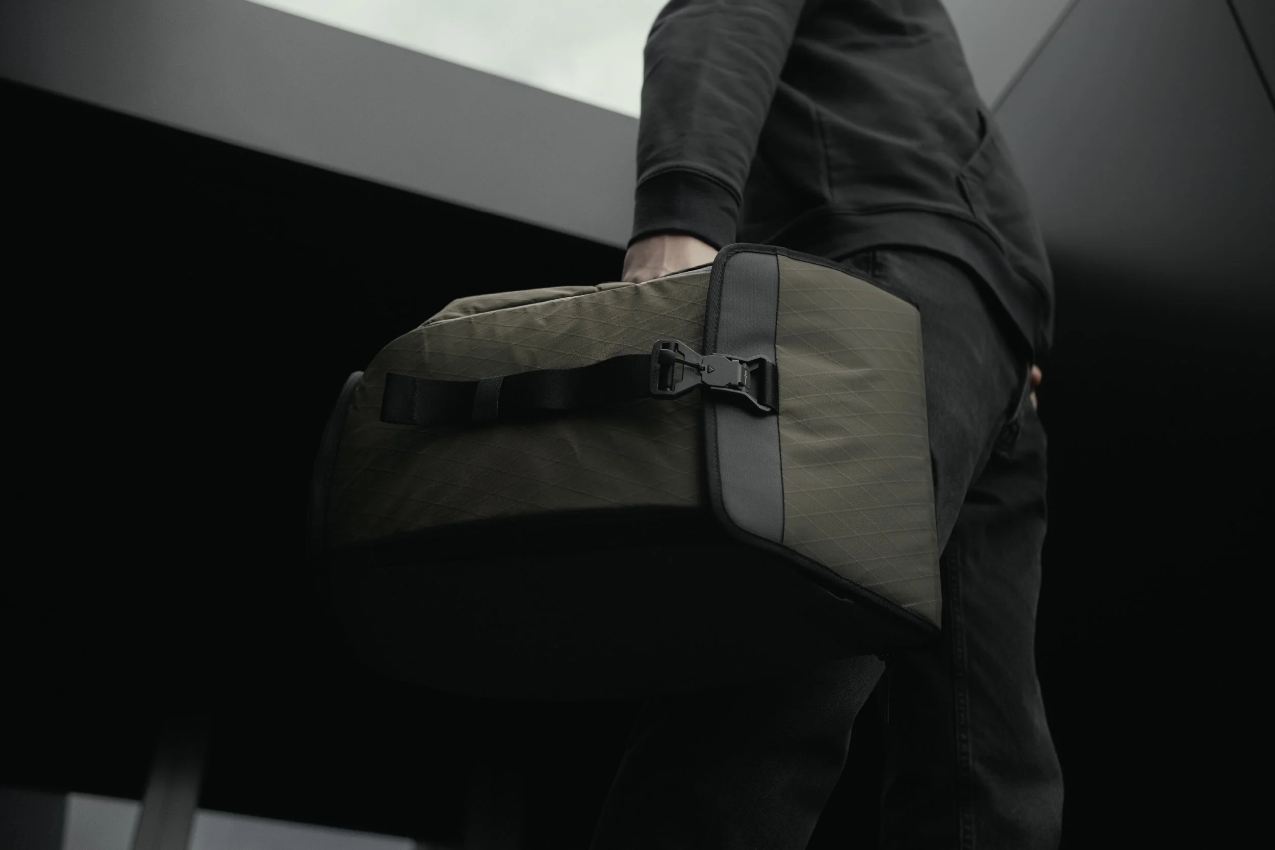 Person in black jacket carrying a green quilted duffel bag with black strap and buckle, standing in a modern indoor setting.