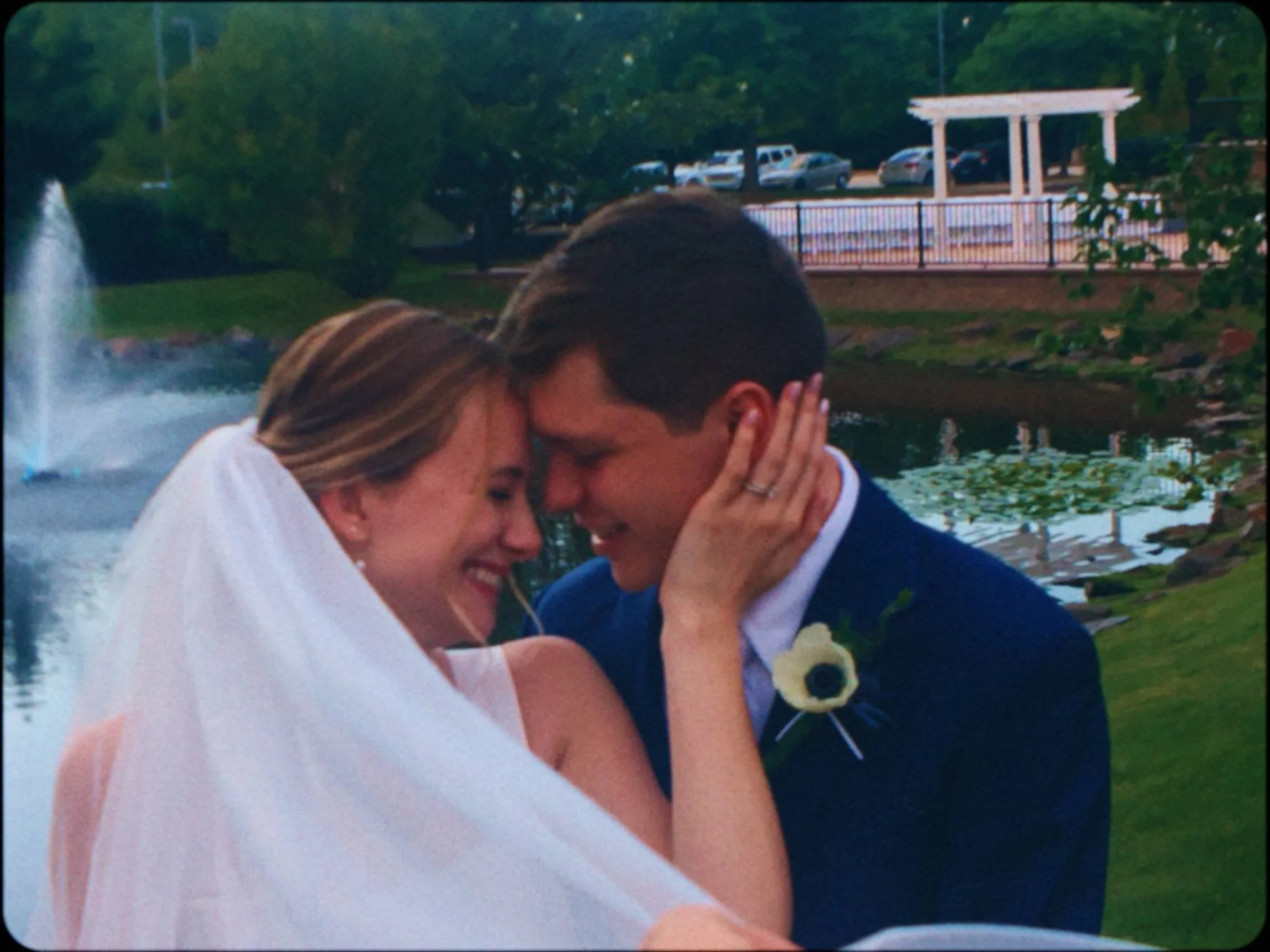 A couple sharing a kiss outdoors near a pond with a fountain. The woman has long hair and is wearing a white dress, and the man has short dark hair and is dressed in a suit. They appear to be celebrating a special occasion, possibly a wedding.