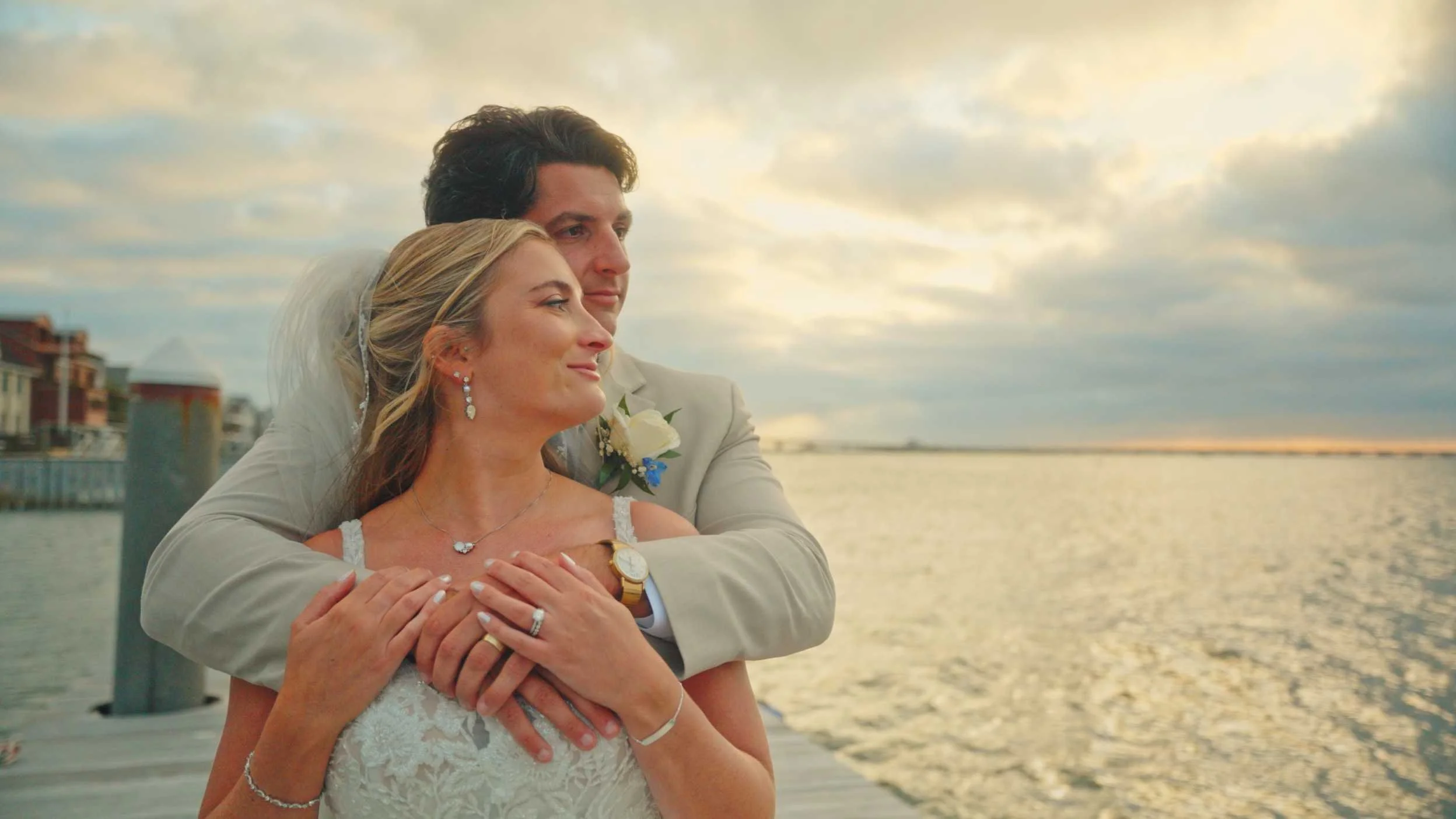 A bride and groom embracing on a dock by the water during sunset, with water and buildings in the background.