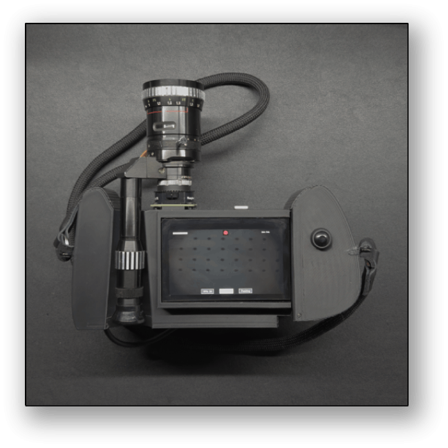 A black camera with a large lens mounted on a support, connected to a rectangular electronic device with a screen and two control buttons, all placed on a black surface.