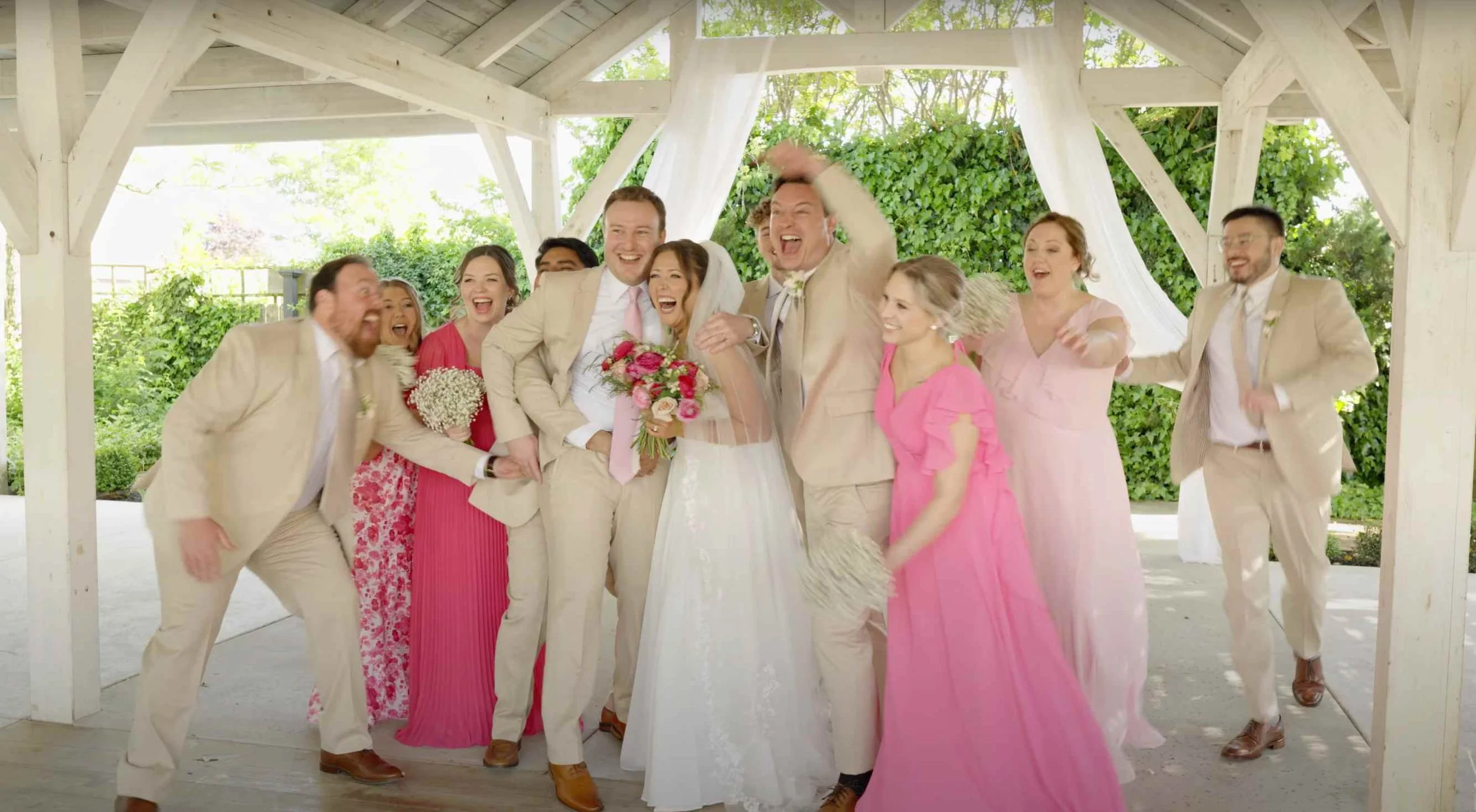 A group of people celebrating at a wedding under a wooden pavilion, wearing light beige suits and pink dresses, with one bride holding a bouquet of pink and white flowers, all smiling and joyful.