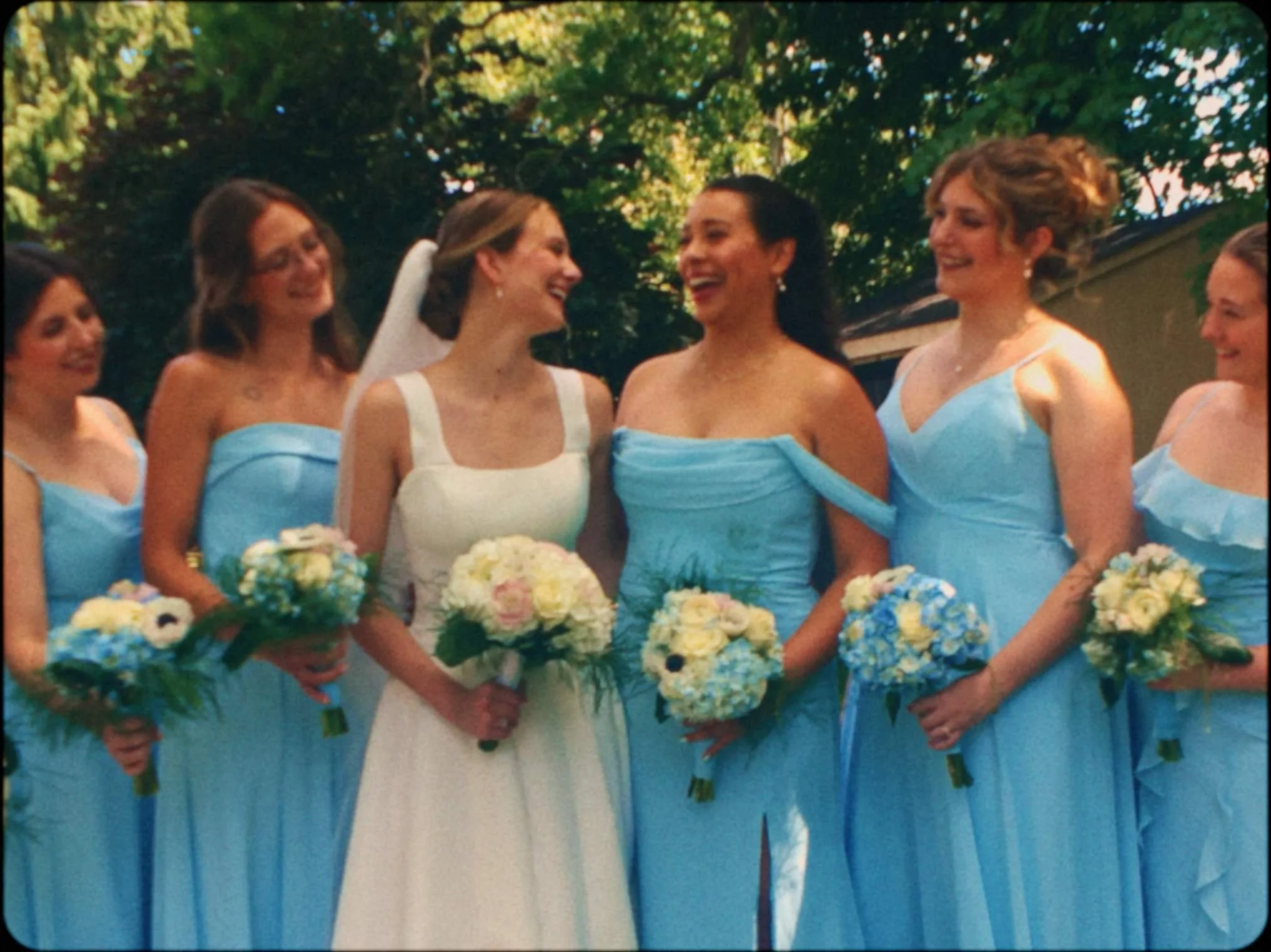 Group of women standing outdoors, dressed in light blue and white dresses, holding bouquets of flowers, smiling and engaging in conversation, with trees in the background.