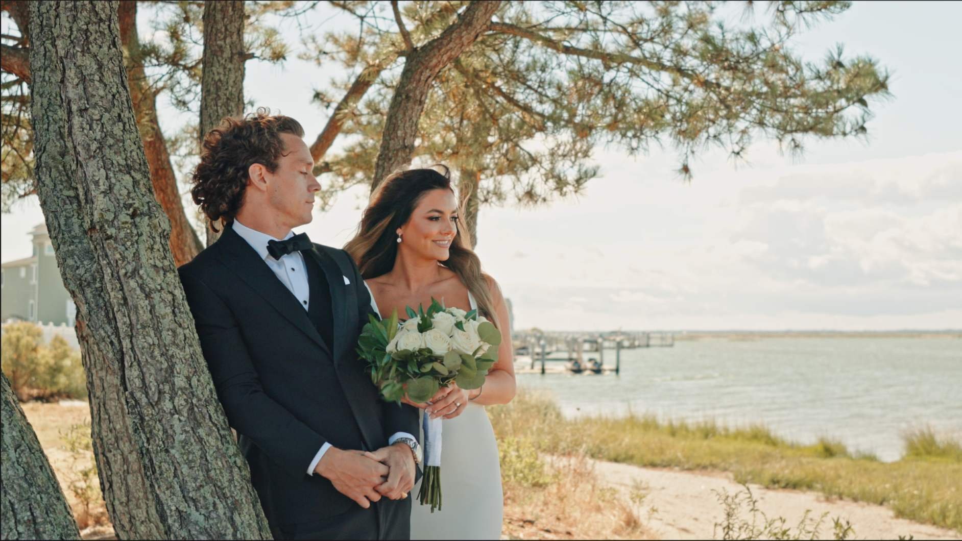 A bride in a white dress holding a bouquet of white roses standing next to a groom in a tuxedo with a black bow tie, both standing near a tree by a body of water with a dock in the background and a cloudy sky.