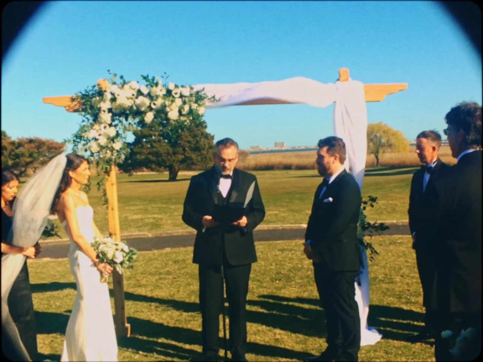 A wedding ceremony outdoors with a cross decorated with flowers and white fabric, where a bride in a white dress and veil holds a bouquet, standing next to bridesmaids, and groomsmen in suits are present with an officiant reading during sunset.