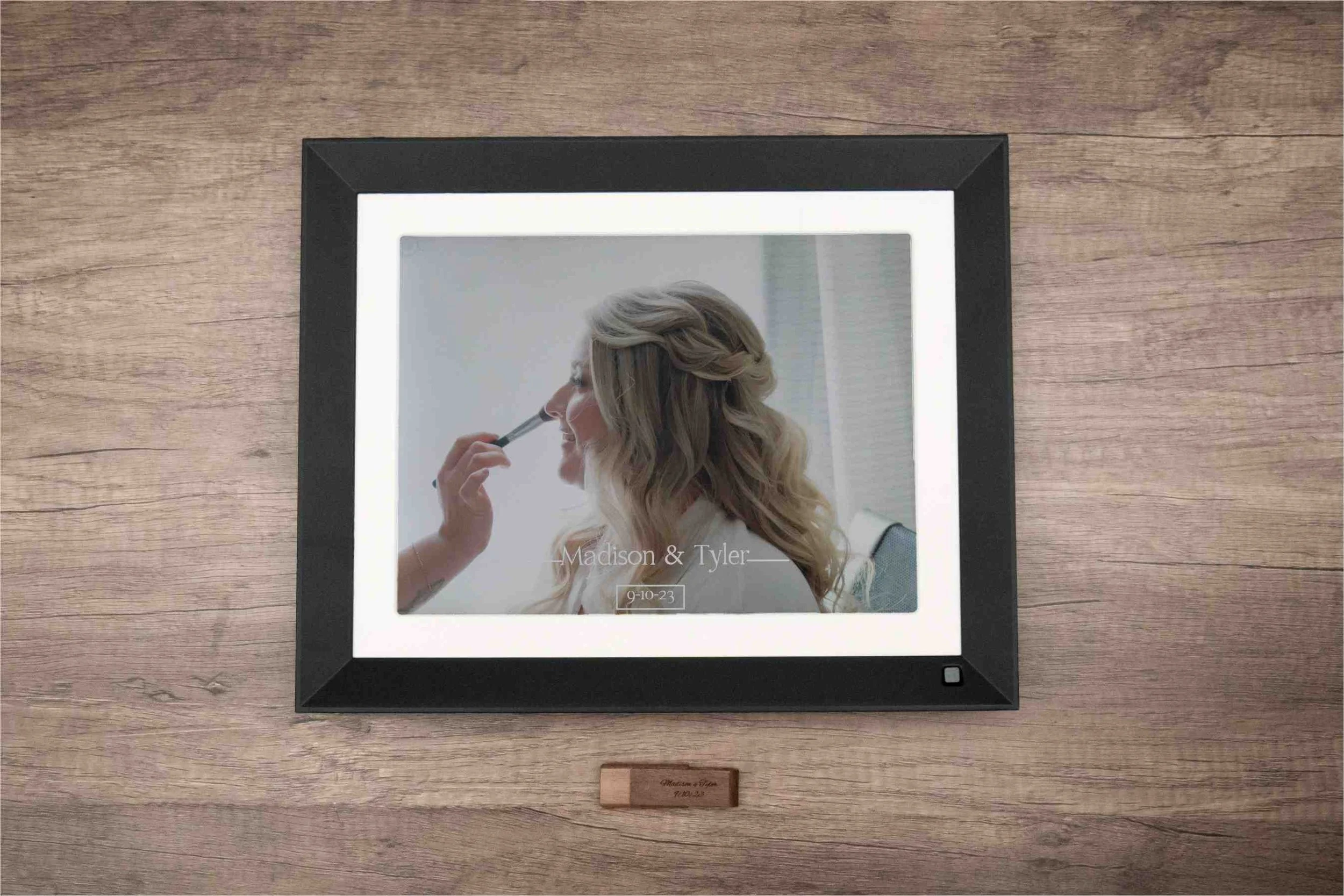 A framed digital picture of a woman with blonde hair applying makeup, with text reading 'Madison & Tyler' and a date '9-10-23' displayed on the screen, mounted on a wooden wall.