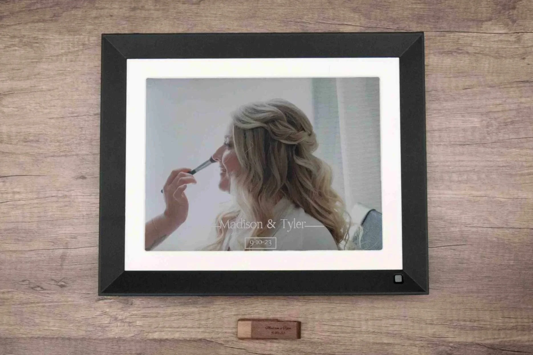 A framed wedding photo on a wooden table showing a woman with blonde hair styled in loose waves and a braid, holding a pen near her nose, with text indicating the names Madison and Tyler and the date 9-10-23.