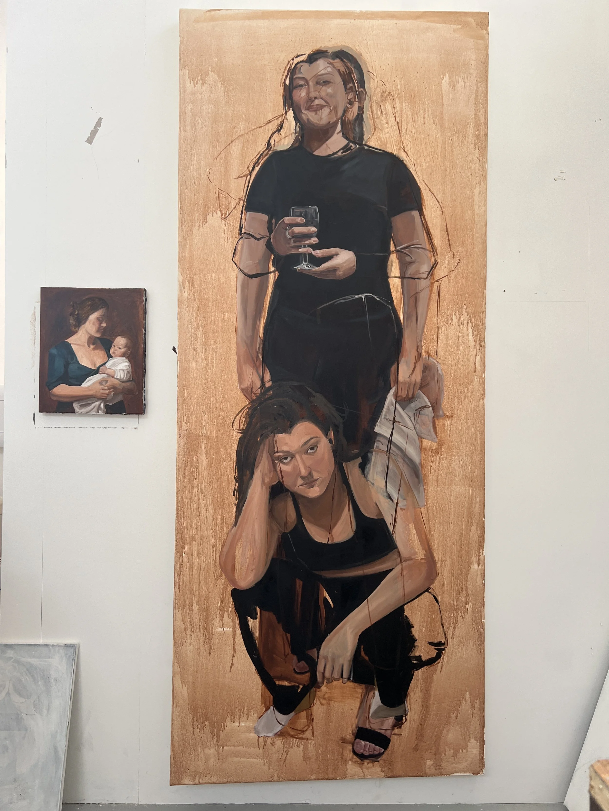 A large painting of two women, one standing and holding a glass of wine and the other sitting, with a smaller portrait of a woman holding a baby on the wall beside it.