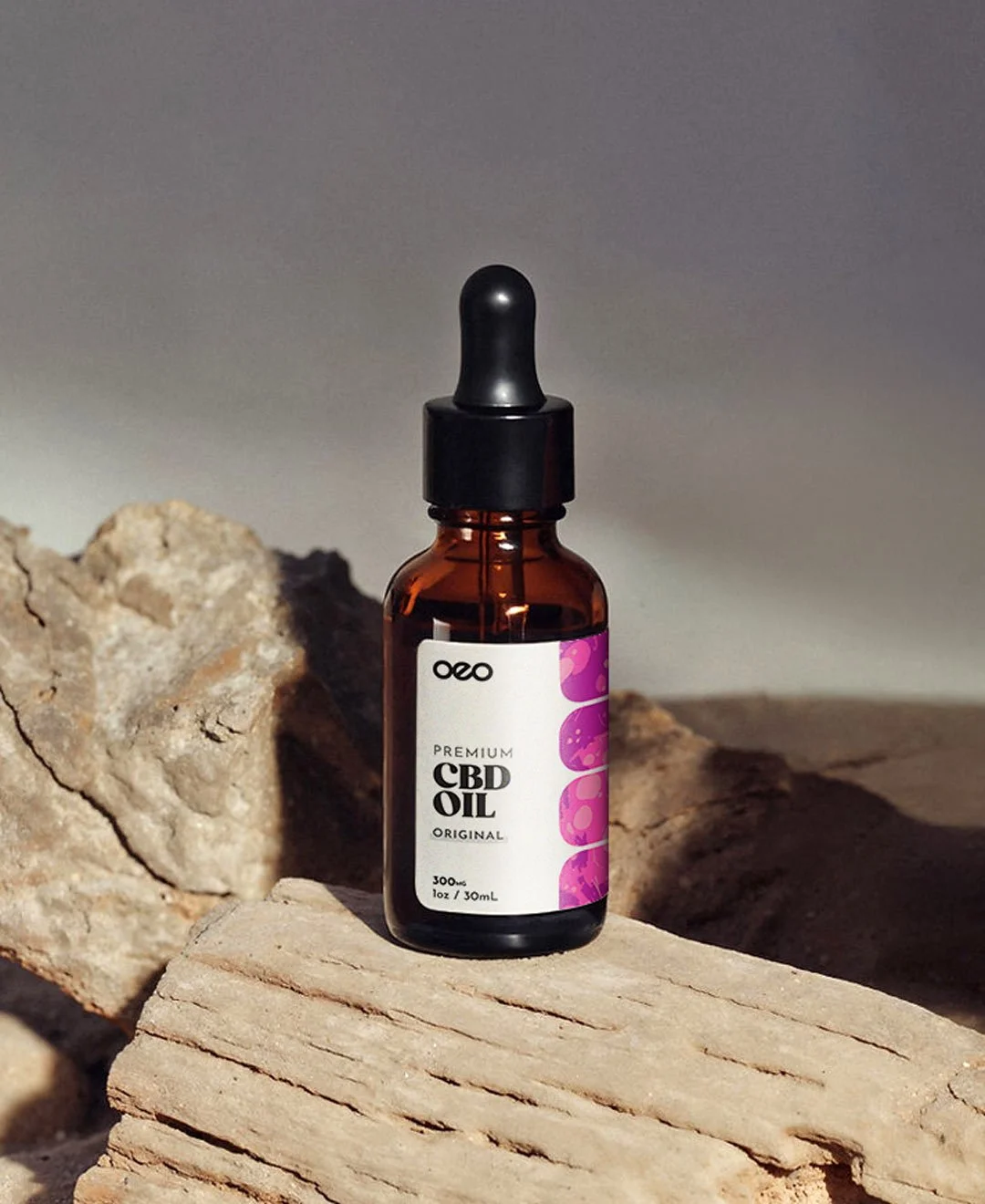 A small amber glass dropper bottle labeled 'Premium CBD Oil' from the brand 'oxo', set on a wooden surface with rocks around, with a plain gradient background.